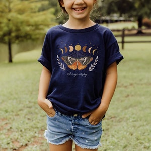 Peut inclure: Un t-shirt bleu marine avec un motif de papillon et le texte "just magic everyday". Le papillon a des ailes orange, brunes et blanches. Le motif comprend &eacute;galement une lune croissante, des &eacute;toiles et un soleil.