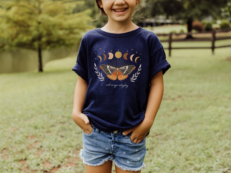 Peut inclure: Un t-shirt bleu marine avec un motif de papillon et le texte "just magic everyday". Le papillon a des ailes orange, brunes et blanches. Le motif comprend &eacute;galement une lune croissante, des &eacute;toiles et un soleil.