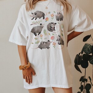May include: White oversized t-shirt with a graphic print of multiple possums in a whimsical floral pattern.