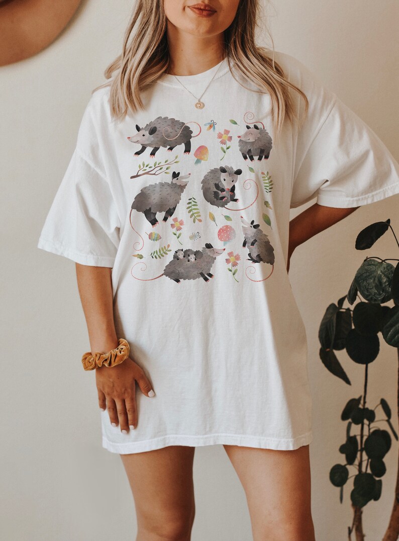May include: White oversized t-shirt with a graphic print of multiple possums in a whimsical floral pattern.