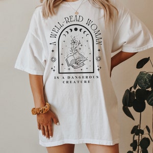 May include: White t-shirt with a black graphic design that reads "A well-read woman is a dangerous creature". The graphic features a book with a flower growing from it, surrounded by stars and a crescent moon.