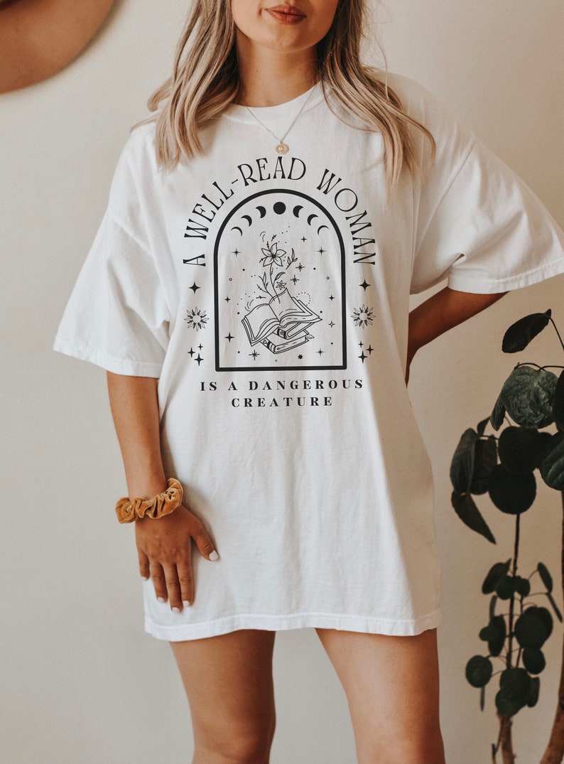 May include: White t-shirt with a black graphic design that reads "A well-read woman is a dangerous creature". The graphic features a book with a flower growing from it, surrounded by stars and a crescent moon.
