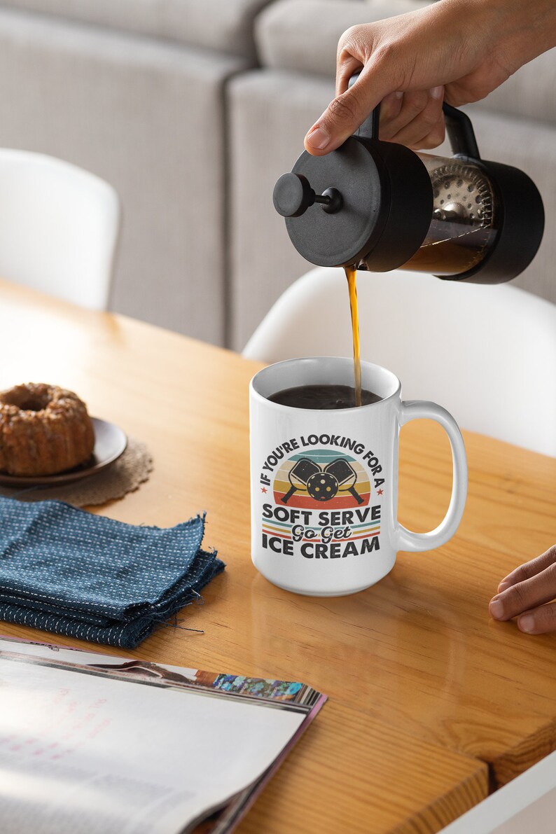 May include: White coffee mug with a retro-style design featuring two pickleball paddles and the text "If you're looking for a soft serve, go get ice cream".