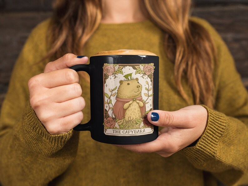 May include: Black ceramic mug with a white illustration of a capybara wearing a crown and a red cape. The illustration is surrounded by a floral border and the text "The Capybara" is printed below the image.