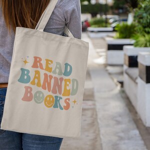 Peut inclure: Un sac fourre-tout en toile blanche avec le texte "READ BANNED BOOKS" en police r&eacute;tro color&eacute;e. Le texte est entour&eacute; d'&eacute;toiles et de visages souriants.