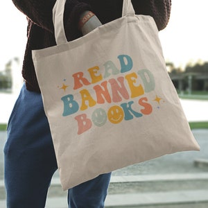 Peut inclure: Un sac fourre-tout beige avec le texte "READ BANNED BOOKS" en couleurs arc-en-ciel avec des smileys.