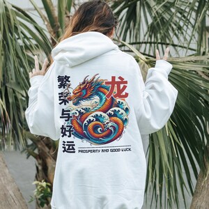 May include: White hoodie with a colorful dragon graphic on the back. The dragon is surrounded by waves and the text "Prosperity and Good Luck" is printed below. The hoodie also features Chinese characters in red.