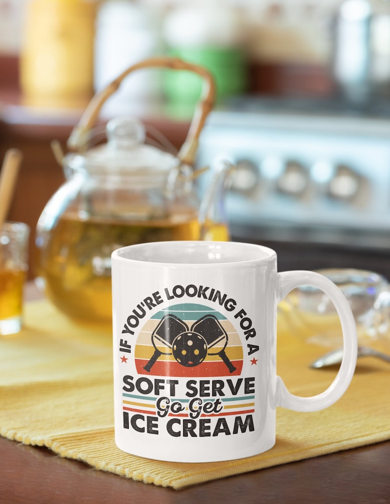 May include: White ceramic mug with a retro-style design featuring two pickleball paddles and a ball. The text reads "If you're looking for a soft serve, go get ice cream."