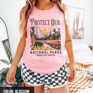May include: Pink tank top with a graphic that reads "PROTECT OUR NATIONAL PARKS" featuring a bear, mountains, and a river. The shorts are black and white checkered with a white trim. The color of the tank top is blossom.