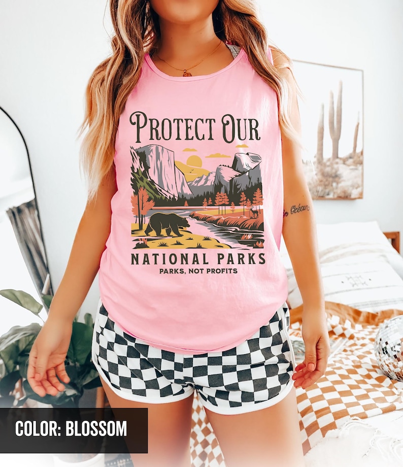 May include: Pink tank top with a graphic that reads "PROTECT OUR NATIONAL PARKS" featuring a bear, mountains, and a river. The shorts are black and white checkered with a white trim. The color of the tank top is blossom.