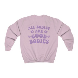 May include: A pale purple sweatshirt featuring the slogan "ALL BODIES ARE GOOD BODIES" in lavender. The design includes flower and butterfly motifs, and two smiley faces. The sweatshirt has a crew neck and long sleeves.