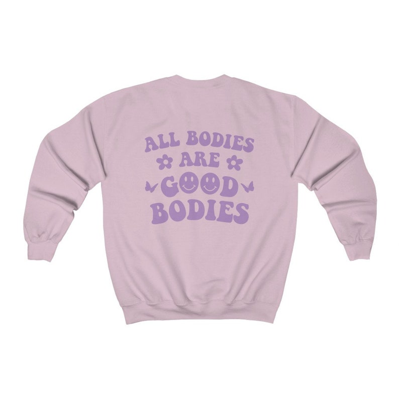 May include: A pale purple sweatshirt featuring the slogan "ALL BODIES ARE GOOD BODIES" in lavender. The design includes flower and butterfly motifs, and two smiley faces. The sweatshirt has a crew neck and long sleeves.