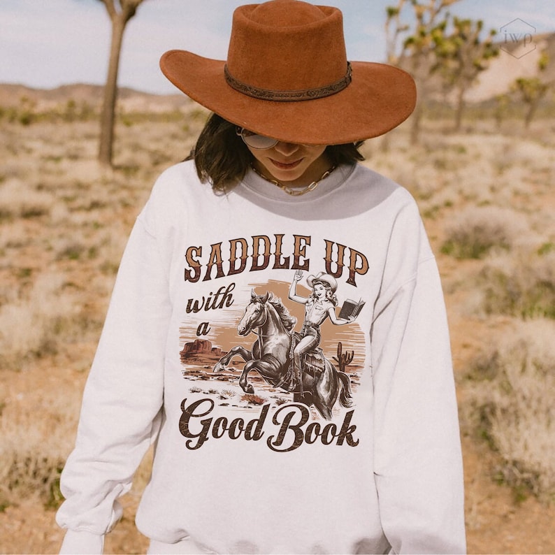 May include: White sweatshirt with a brown graphic print of a woman riding a horse and holding a book. The text "Saddle Up with a Good Book" is printed below the graphic.