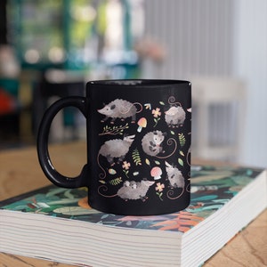 May include: Black ceramic mug with a white handle. The mug features a repeating pattern of opossums, mushrooms, and flowers on a black background.