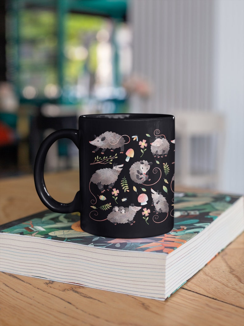 May include: Black ceramic mug with a white handle. The mug features a repeating pattern of opossums, mushrooms, and flowers on a black background.