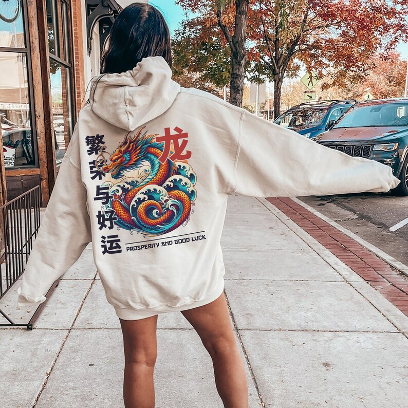 May include: A cream-colored hoodie with a large graphic print of a red dragon with blue and orange waves. The text "Prosperity and Good Luck" is printed below the dragon. The Chinese characters "繁荣与好运" are printed on the left side of the dragon.