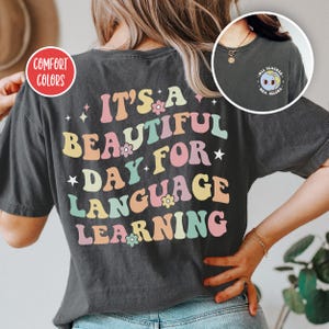 May include: A charcoal gray t-shirt featuring the text IT'S A BEAUTIFUL DAY FOR LANGUAGE LEARNING in pastel pink, yellow, and teal wavy lettering with small flower and star accents. The front chest area displays a small globe graphic with the text MLL TEACHER MRS. SELENA.
