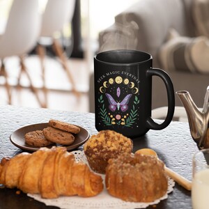 May include: Black ceramic mug with a white design featuring a moth, crystals, and the text "Seek Magic Everyday". The mug is sitting on a table with pastries and cookies.