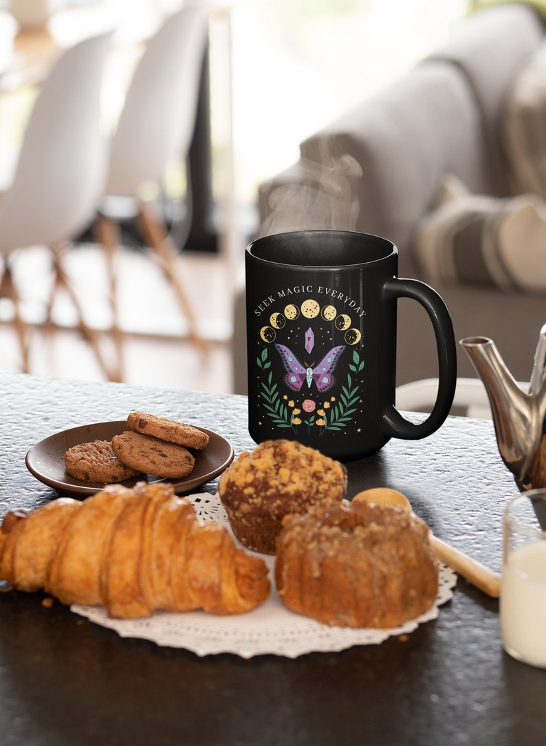 May include: Black ceramic mug with a white design featuring a moth, crystals, and the text "Seek Magic Everyday". The mug is sitting on a table with pastries and cookies.