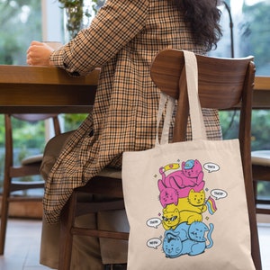 K&ouml;nnte beinhalten: Eine beigefarbene Tragetasche aus Segeltuch mit einer grafischen Illustration von vier gestapelten Katzen in Pink, Gelb und Blau. Die Katzen tragen Accessoires wie eine blaue M&uuml;tze und eine Sonnenbrille. Sprechblasen enthalten den Text: They, their, them, meow.