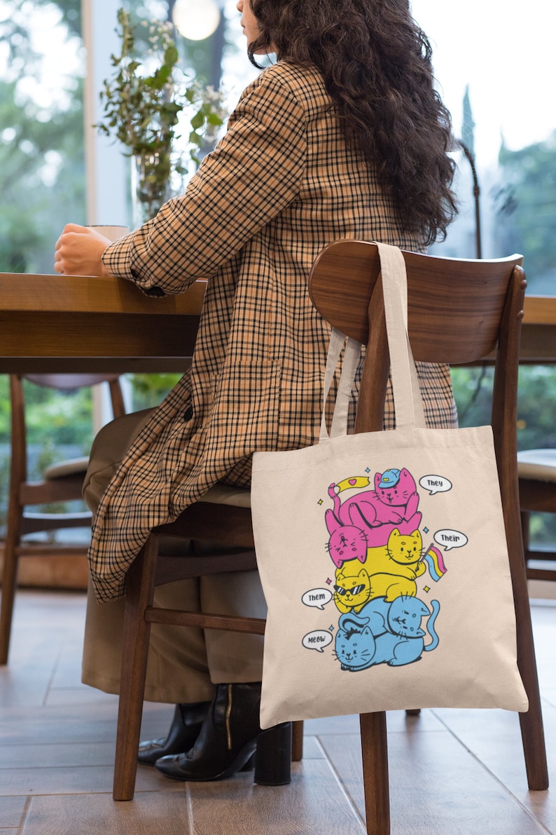 K&ouml;nnte beinhalten: Eine beigefarbene Tragetasche aus Segeltuch mit einer grafischen Illustration von vier gestapelten Katzen in Pink, Gelb und Blau. Die Katzen tragen Accessoires wie eine blaue M&uuml;tze und eine Sonnenbrille. Sprechblasen enthalten den Text: They, their, them, meow.