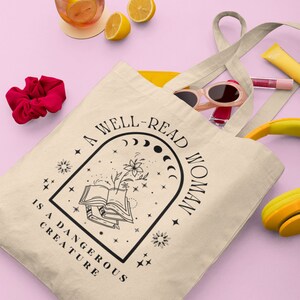 Pode incluir: Bolsa tote bege com texto preto que diz "A well-read woman is a dangerous creature". A bolsa cont&eacute;m &oacute;culos de sol, brilho labial e uma carteira amarela. Uma bebida com rodelas de lim&atilde;o e uma presilha de cabelo vermelha tamb&eacute;m s&atilde;o vis&iacute;veis.