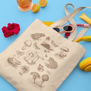 May include: A beige canvas tote bag with a brown illustration of mushrooms, frogs, butterflies, and other forest elements. The bag has a yellow lining and is partially filled with sunglasses, a pink lip gloss, and a yellow headband.