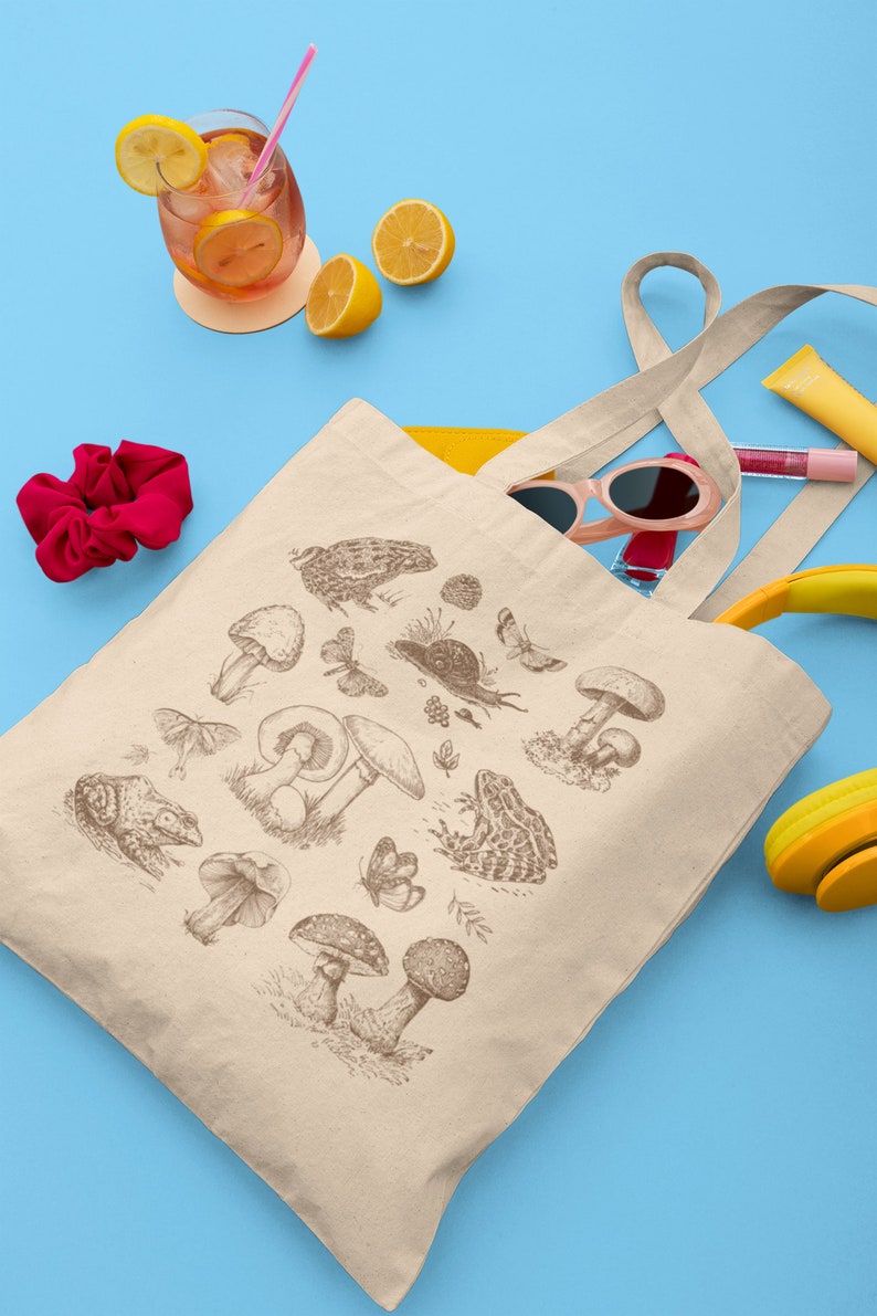 May include: A beige canvas tote bag with a brown illustration of mushrooms, frogs, butterflies, and other forest elements. The bag has a yellow lining and is partially filled with sunglasses, a pink lip gloss, and a yellow headband.