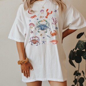 May include: White oversized t-shirt with a colorful watercolor design of various sea creatures, including crabs, lobsters, starfish, and seashells.