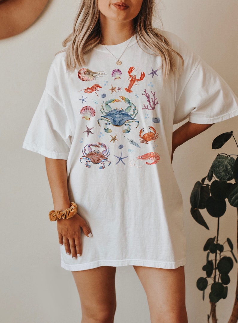 May include: White oversized t-shirt with a colorful watercolor design of various sea creatures, including crabs, lobsters, starfish, and seashells.