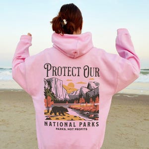 May include: A light pink hoodie with a graphic on the back that says "Protect Our National Parks." The graphic features a bear, mountains, and a river. The text "Parks, Not Profits" is below the graphic.
