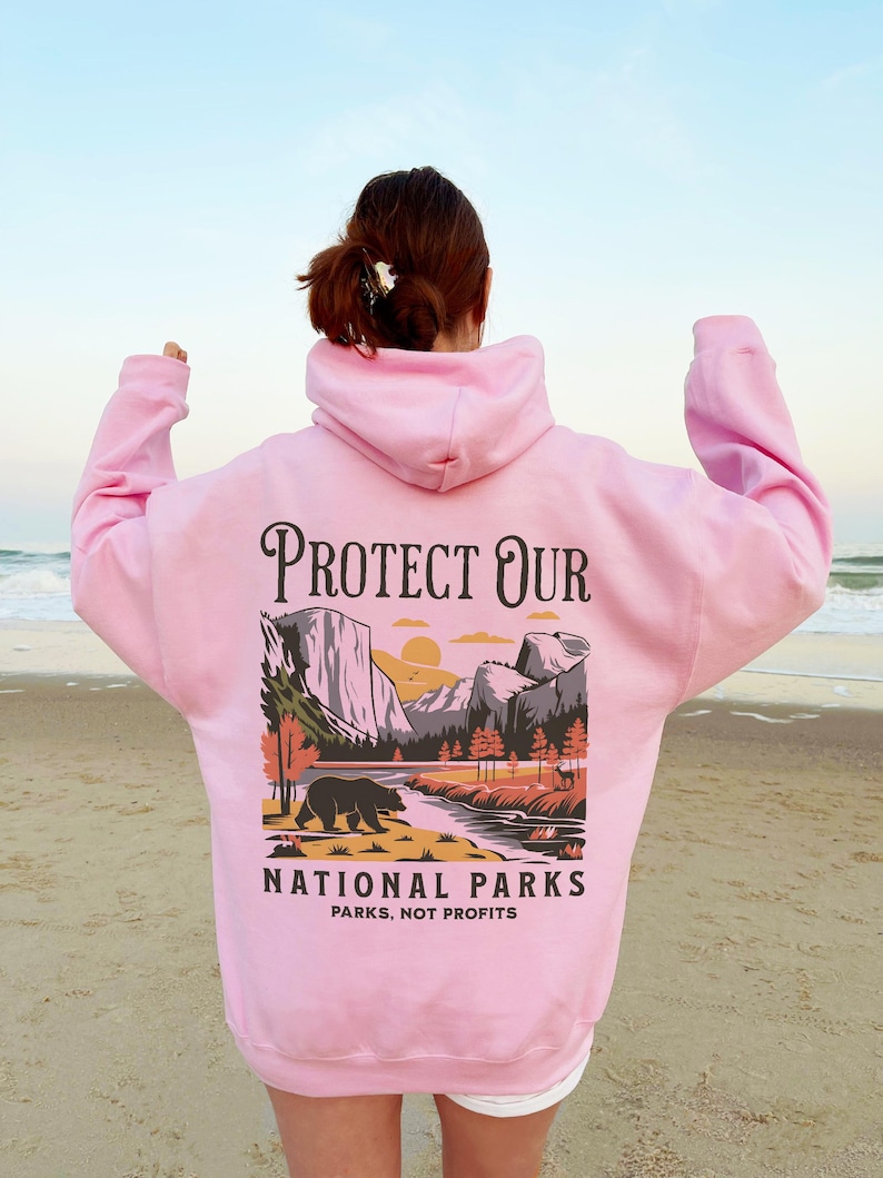 May include: A light pink hoodie with a graphic on the back that says "Protect Our National Parks." The graphic features a bear, mountains, and a river. The text "Parks, Not Profits" is below the graphic.