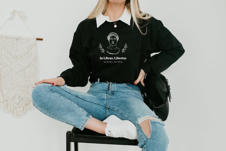 May include: A black sweatshirt with a white graphic of a woman's head with the text "In Libras, Libertas" and "In books, freedom" underneath. The model is wearing blue jeans and white sneakers.