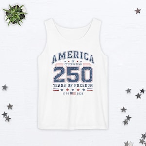 以下が含まれることがあります： 「AMERICA CELEBRATING 250 YEARS OF FREEDOM」と1776年と2026年の文字が入った、青と赤のグラフィックが施された白いタンクトップ。デザインには星とストライプが含まれています。