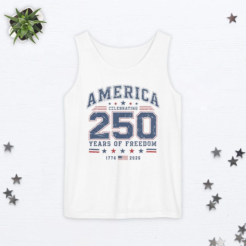 以下が含まれることがあります： 「AMERICA CELEBRATING 250 YEARS OF FREEDOM」と1776年と2026年の文字が入った、青と赤のグラフィックが施された白いタンクトップ。デザインには星とストライプが含まれています。