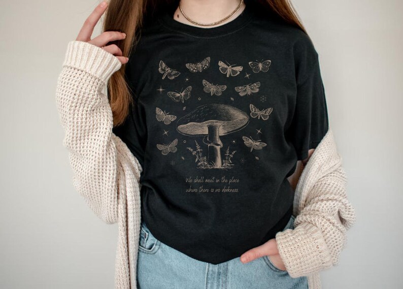 May include: Black t-shirt with a mushroom and butterflies graphic. The text "We shall meet in the place where there is no darkness" is printed on the shirt.