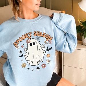 May include: A light blue sweatshirt with a graphic of a ghost with the text "Spooky Season" in a retro font. The ghost is wearing a peace sign necklace and has flowers and stars around it.