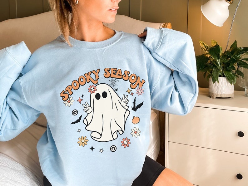 May include: A light blue sweatshirt with a graphic of a ghost with the text "Spooky Season" in a retro font. The ghost is wearing a peace sign necklace and has flowers and stars around it.