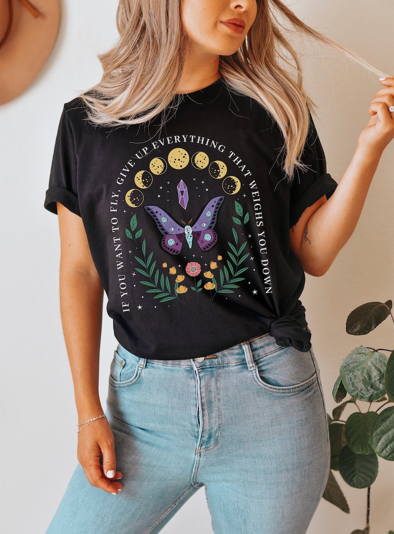 May include: Black t-shirt with a white graphic print of a butterfly, moon phases, and a crystal. The text reads "If you want to fly, give up everything that weighs you down."