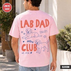 Puede incluir: Una camiseta de manga corta rosa claro con un gr&aacute;fico en la espalda que incluye el texto LAB DAD CLUB, est. 2026, y home is where my labrador is. El dise&ntilde;o muestra dos ilustraciones de Labrador, huellas, una pelota y estrellas.
