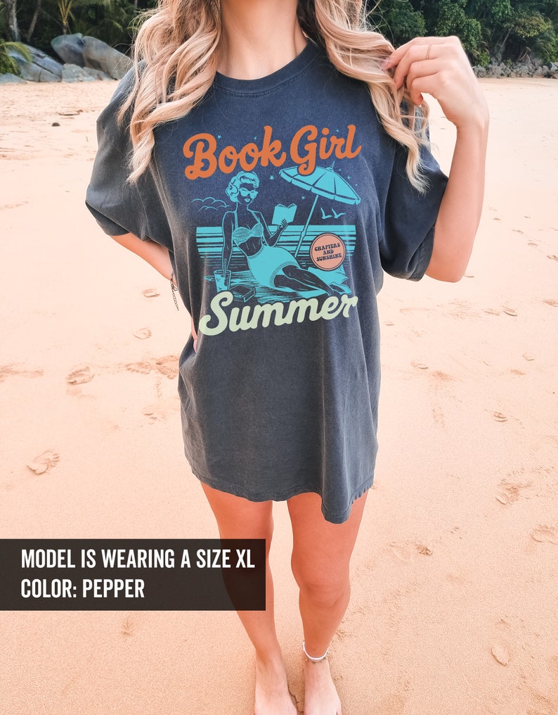 K&ouml;nnte beinhalten: Ein dunkelgraues T-Shirt mit einer Retro-Illustration einer Frau, die am Strand ein Buch liest. Der Text "Book Girl Summer" ist auf dem T-Shirt aufgedruckt. Der Text "Chapters are my sunshine" ist in einem Kreis auf dem T-Shirt aufgedruckt.