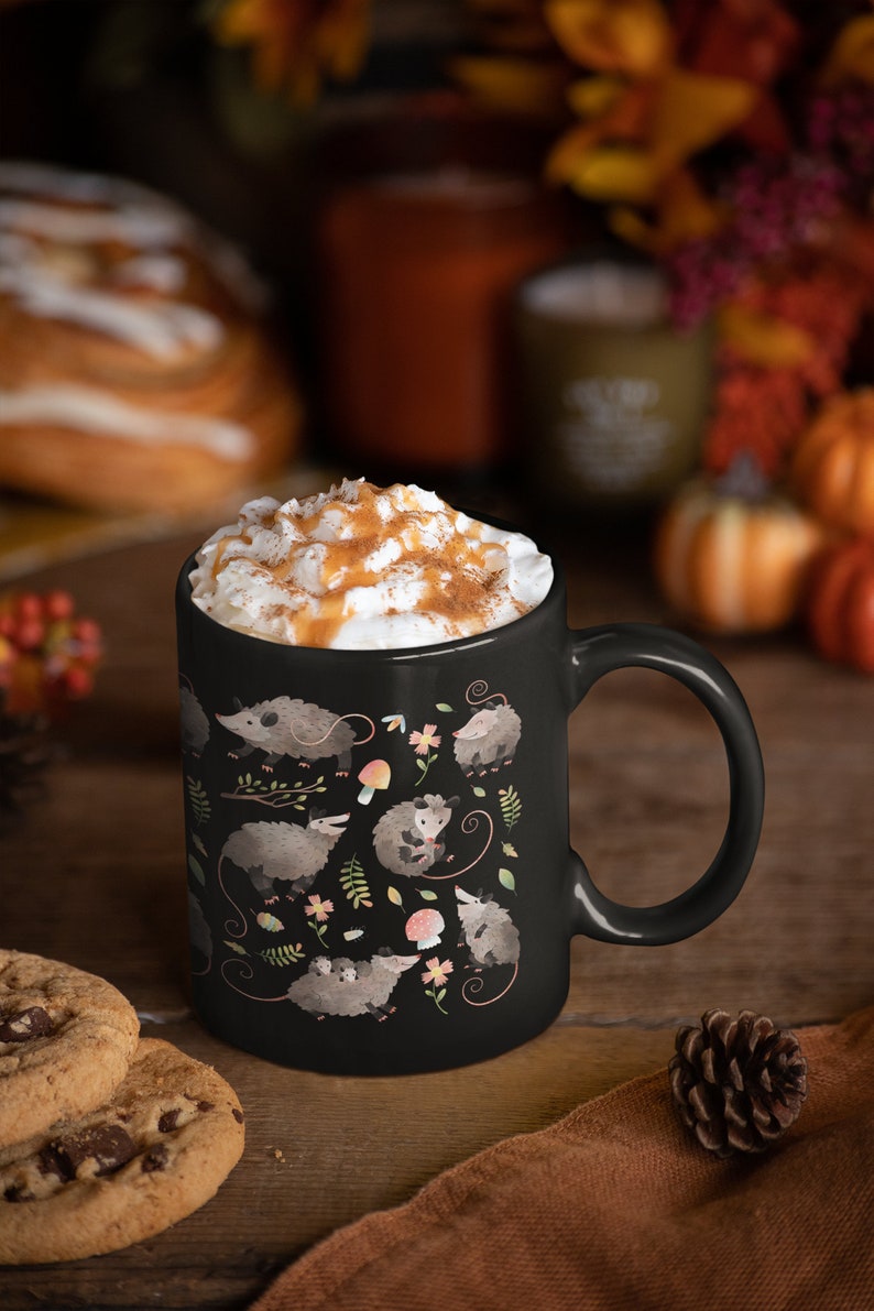 May include: A black ceramic mug with a white rim and handle. The mug is filled with a creamy beverage topped with caramel and cinnamon. The mug features a repeating pattern of opossums and autumn leaves in shades of brown, pink, and green.