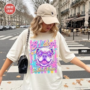 May include: An ivory t-shirt featuring a graphic of a bulldog wearing star-shaped sunglasses, surrounded by colorful stars and vertical stripes. The text reads "Bulldogs" and "School Name". The shirt is paired with a beige baseball cap and a black bag.
