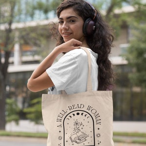 Pode incluir: Saco tote bege com o texto "A WELL-READ WOMAN IS A DANGEROUS CREATURE" em preto. O design inclui um arco, luas, estrelas e um livro aberto com uma flor. O saco &eacute; transportado ao ombro.
