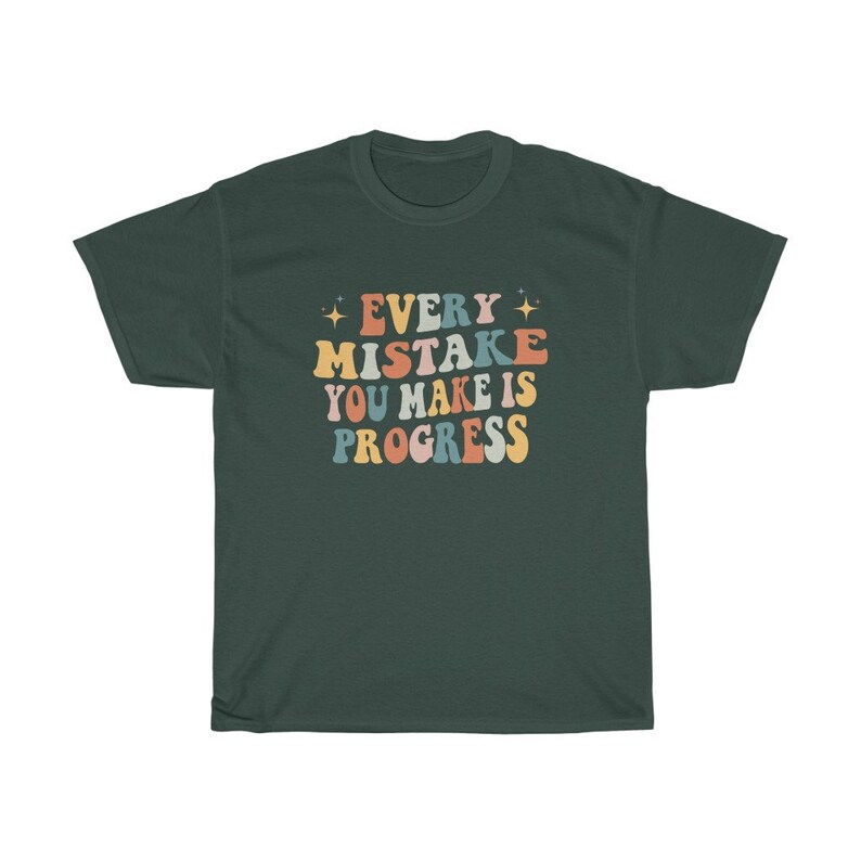 May include: Dark green t-shirt with the text "EVERY MISTAKE YOU MAKE IS PROGRESS" in a retro font. The text is in various colors, including orange, blue, and yellow. The shirt has short sleeves and a crew neck.