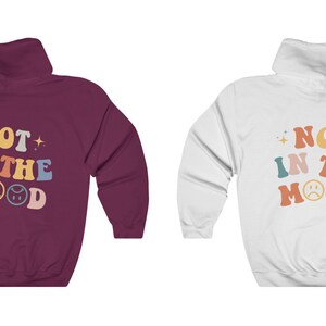 Pu&ograve; includere: Due felpe con cappuccio con il testo "NOT IN THE MOOD" in lettere retr&ograve; colorate e due emoji con facce tristi. Una felpa &egrave; color granata e l'altra &egrave; bianca.
