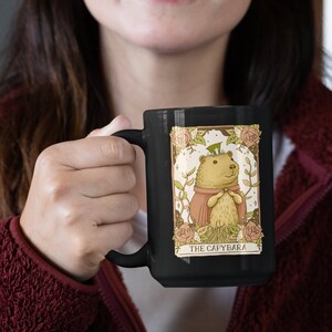 May include: A black ceramic mug with a white design of a capybara wearing a crown and a red cape. The design is surrounded by flowers and stars. The text "THE CAPYBARA" is printed below the image.