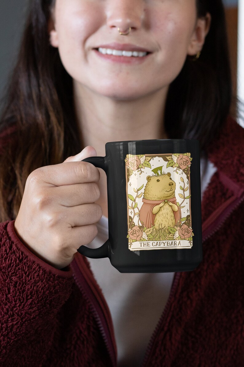 May include: A black ceramic mug with a white design of a capybara wearing a crown and a red cape. The design is surrounded by flowers and stars. The text "THE CAPYBARA" is printed below the image.