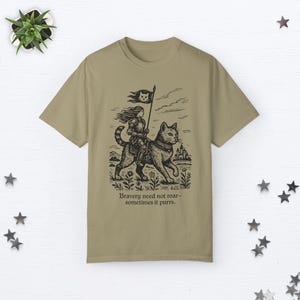 Peut inclure: Un t-shirt kaki avec une illustration en noir et blanc d'une femme chevauchant un chat. La femme porte une armure et tient un drapeau. Le texte "Bravery need not roar - sometimes it purrs." est imprim&eacute; sous l'illustration.