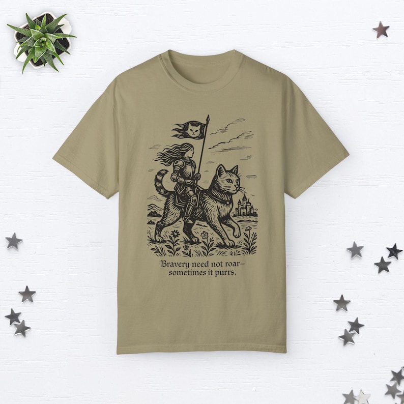 Peut inclure: Un t-shirt kaki avec une illustration en noir et blanc d'une femme chevauchant un chat. La femme porte une armure et tient un drapeau. Le texte "Bravery need not roar - sometimes it purrs." est imprim&eacute; sous l'illustration.